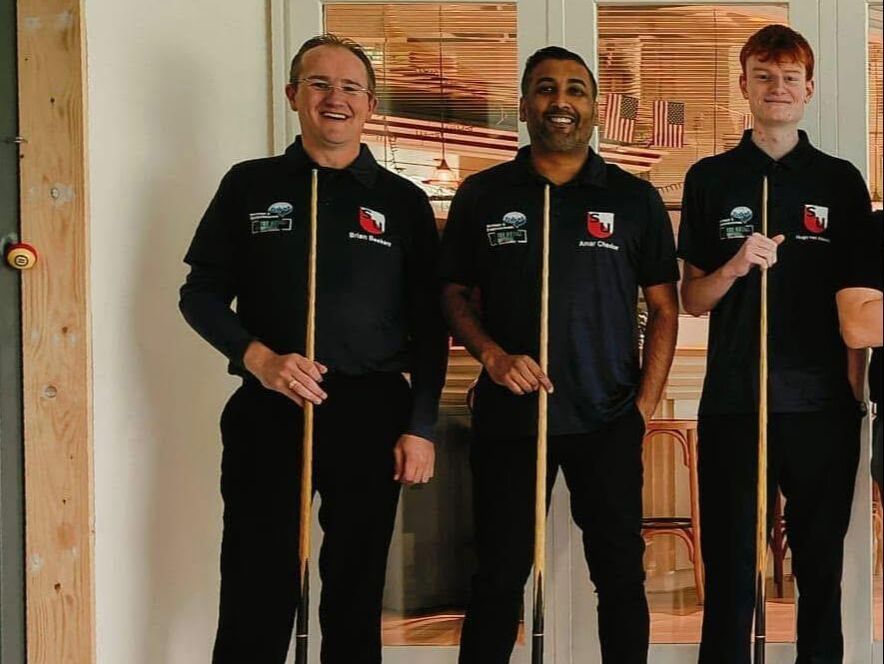 Drie mannen met biljartkeuen poseren glimlachend binnen. Donkere shirts, zwarte broeken.