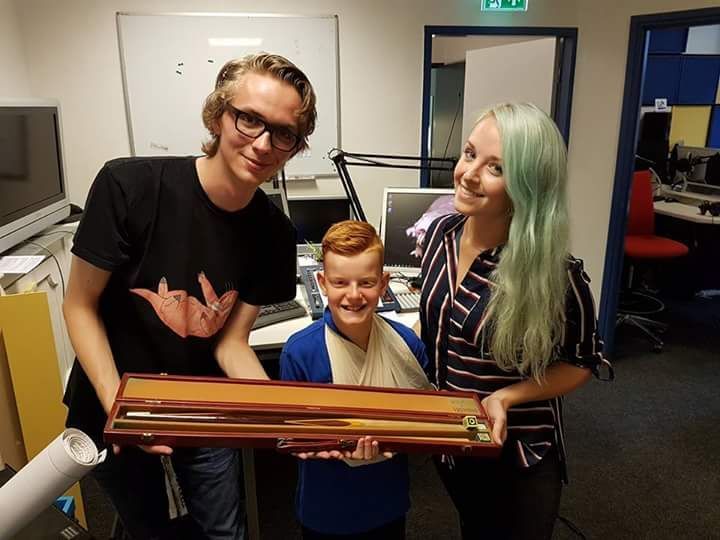 Drie mensen houden een vioolkoffer vast; een jongen met een draagdoek glimlacht tussen twee volwassenen op een kantoor.