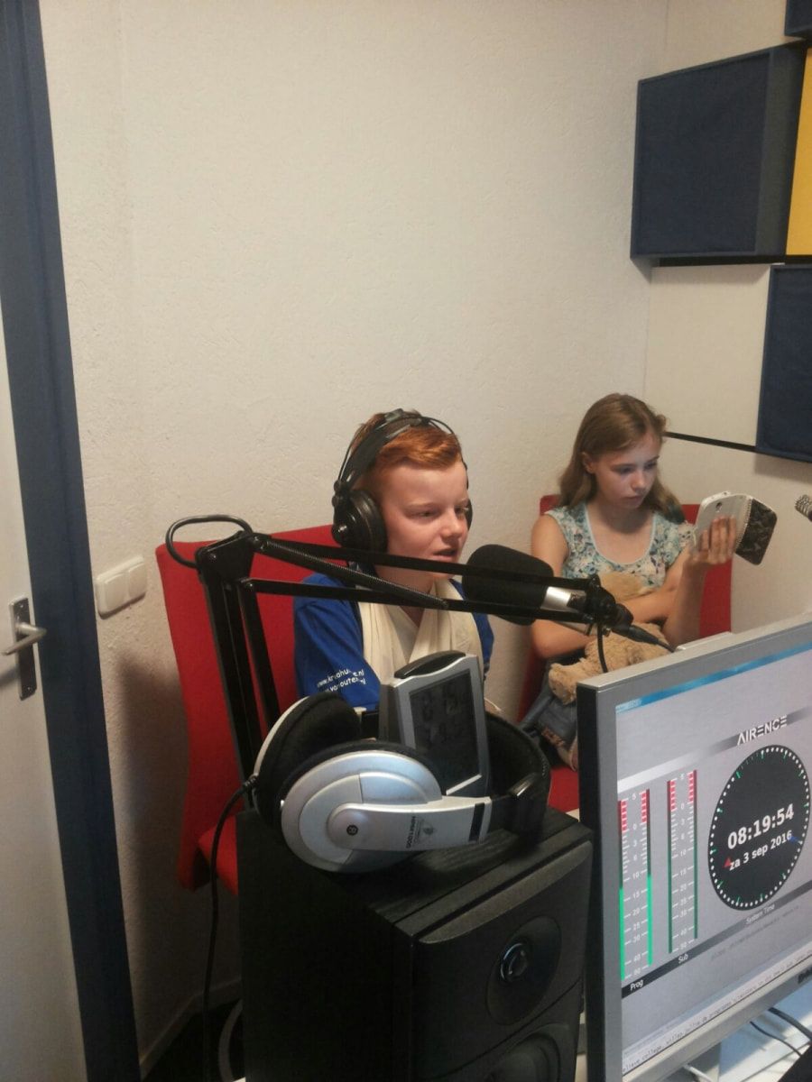 Twee kinderen in een opnamestudio, met koptelefoons op, pratend in microfoons. De een kijkt vooruit; de ander houdt een telefoon vast.
