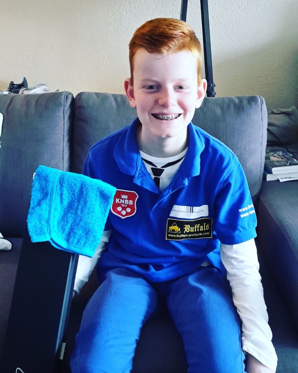 Een jonge jongen met rood haar en een beugel draagt ​​een blauw uniform. Hij lacht en houdt een keu-koffer en een blauwe handdoek vast op een bank.