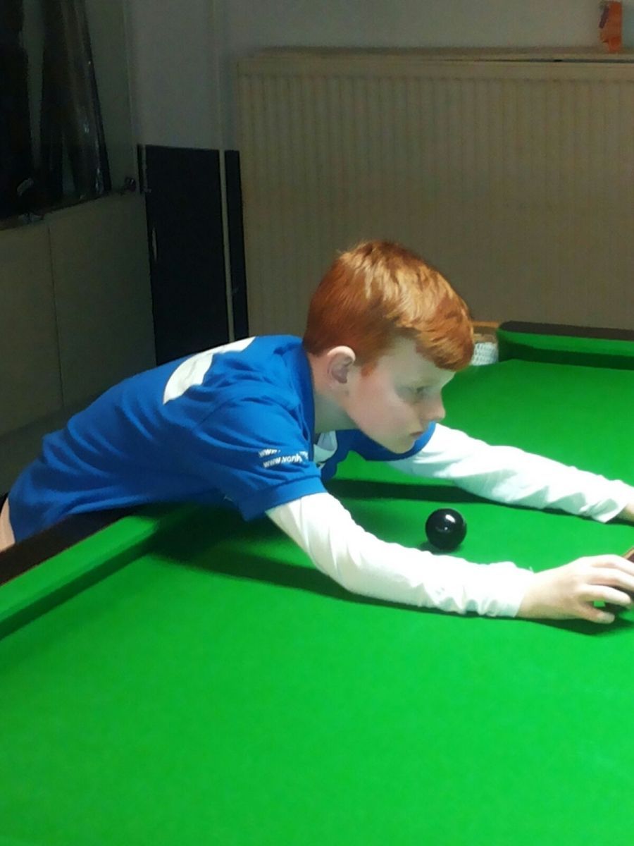 Jongen met rood haar, die biljart speelt, mikt op een zwarte bal op een groene tafel, gekleed in een blauw en wit shirt.