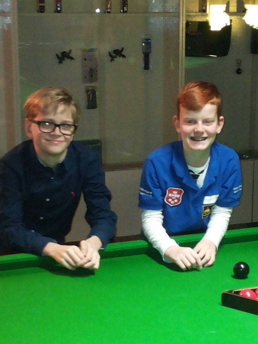 Twee lachende jongens aan een snookertafel. Een met een bril in een donkerblauw shirt, de ander in een blauw uniform.
