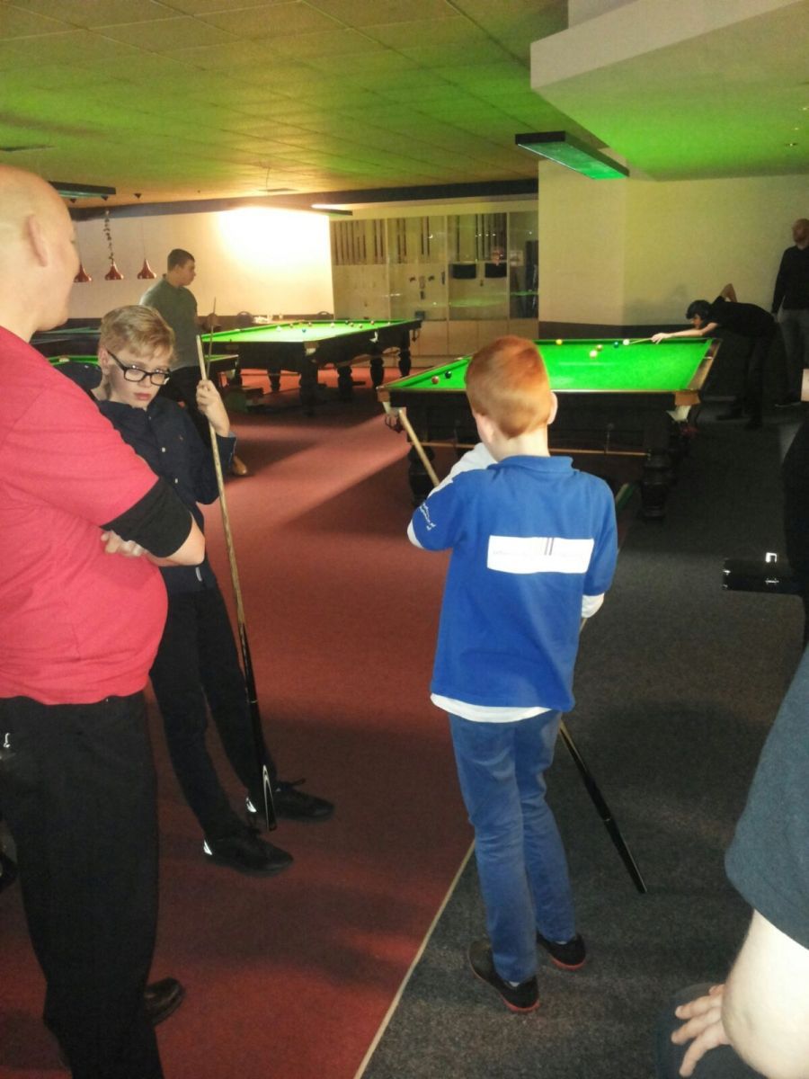 Een groep mensen in een snookerhal; twee jongens en een man kijken toe hoe een andere jongen op een tafel richt.