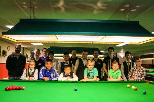 Een groep mensen poseert achter een snookertafel. Spelers van verschillende leeftijden met keuen in een poolzaal.