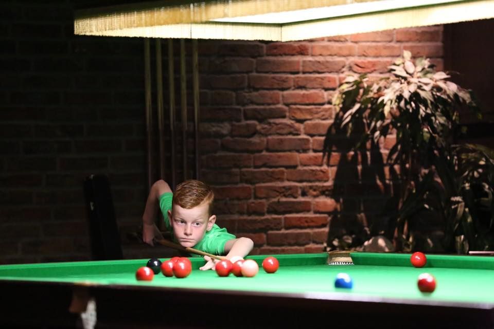 Een jonge jongen richt zijn pijlen op snookerballen op een groene tafel, tegen een achtergrond van een fel licht en een bakstenen muur.