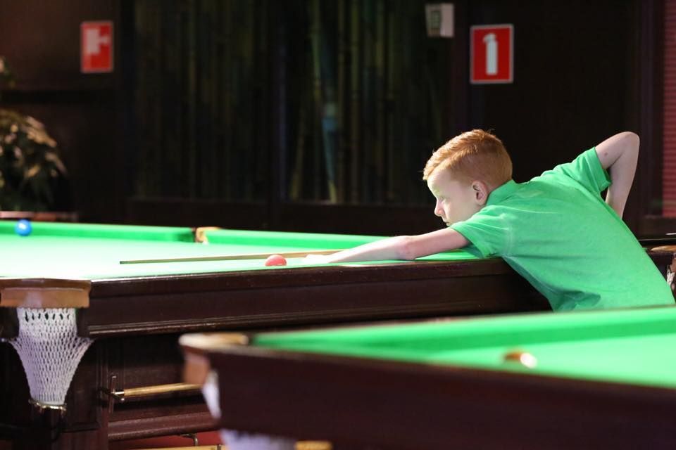 Een jongetje in een groen shirt speelt biljart in een slecht verlichte kamer.