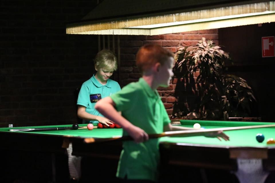 Twee jongens spelen pool in een slecht verlichte kamer. De een richt met een keu, de ander legt ballen neer.