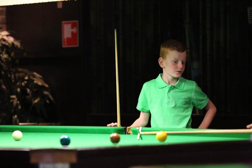 Jongen in een groen shirt speelt biljart in een slecht verlichte kamer, met een keu in de hand, kijkend naar de tafel.