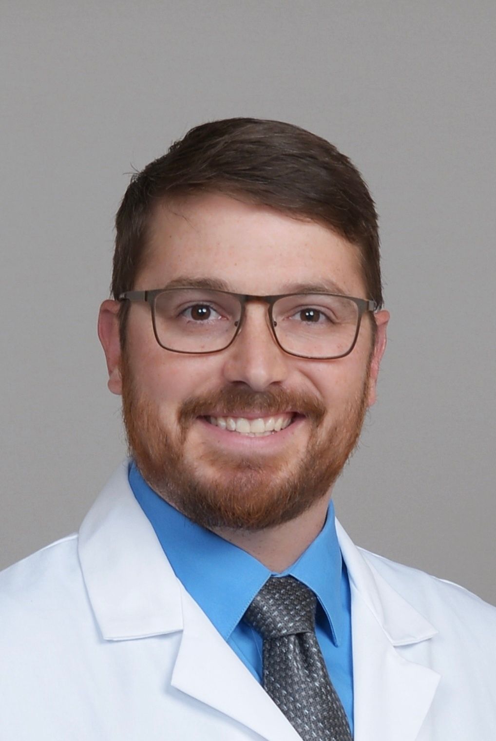 Man in a blue shirt, tie, and gray suit jacket, smiling, headshot against a gray background.
