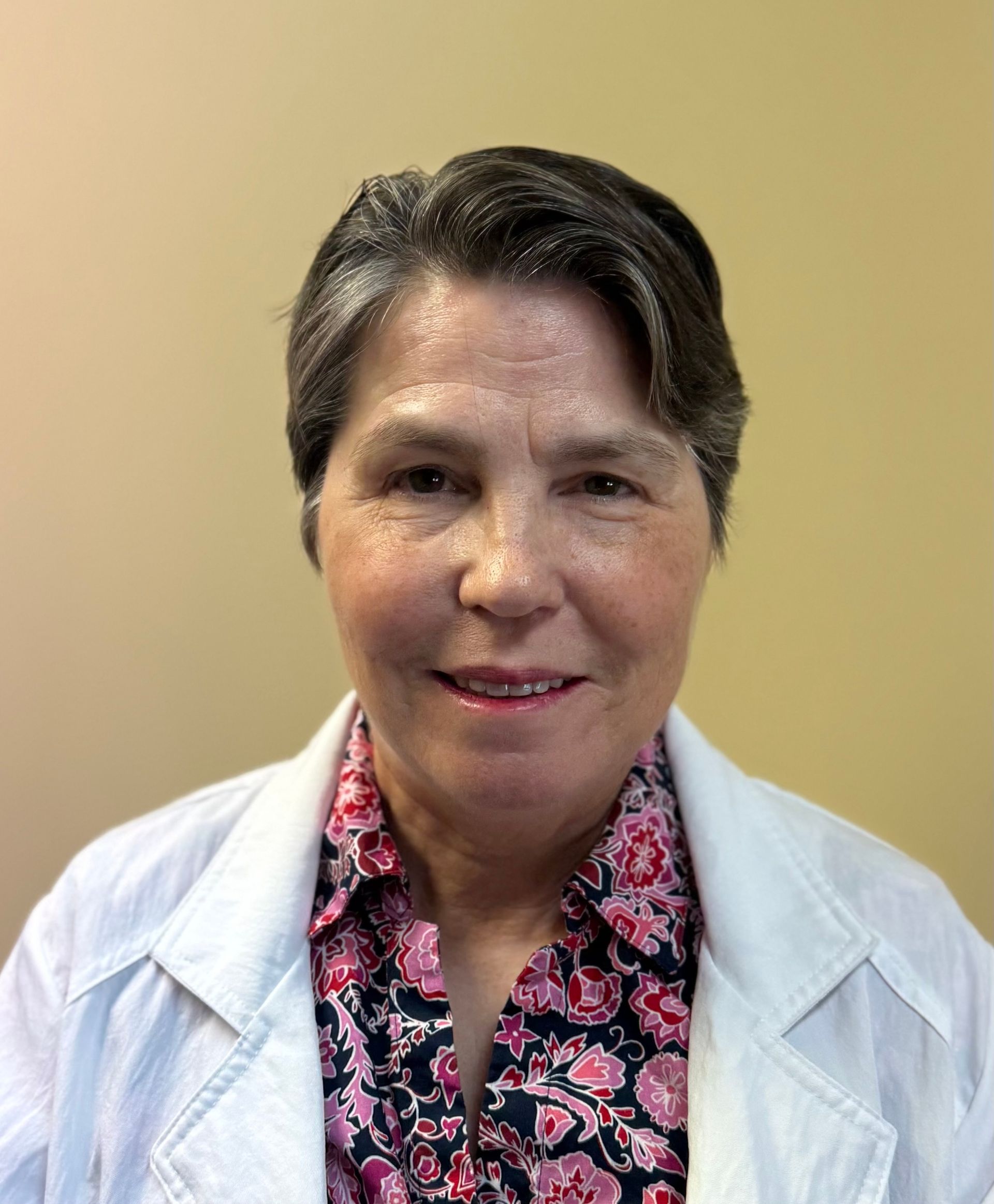 Woman in a white coat and floral shirt smiles, in front of a neutral-colored wall.