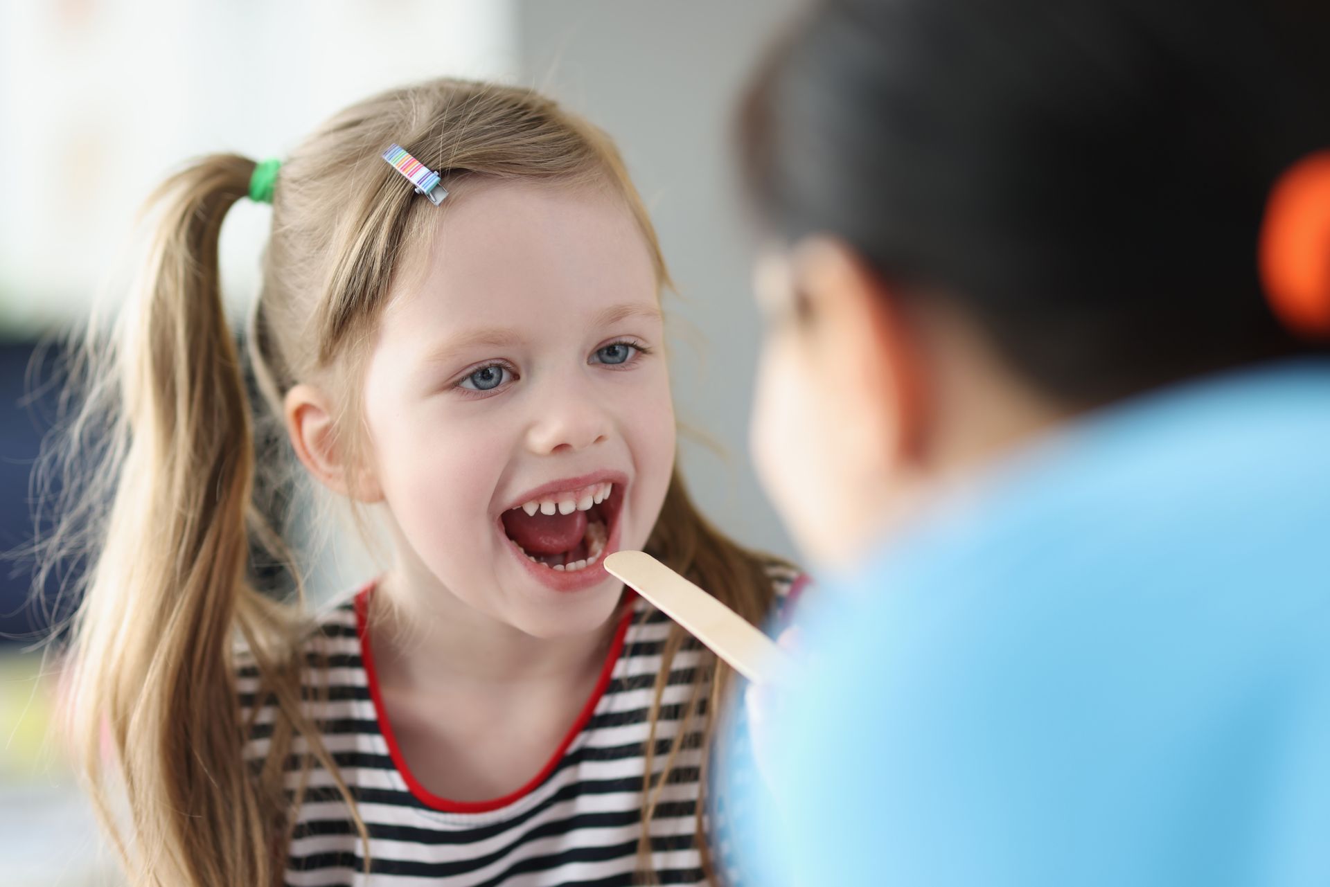 Young child with pigtails has mouth open for examination; a medical professional uses a tongue depressor.