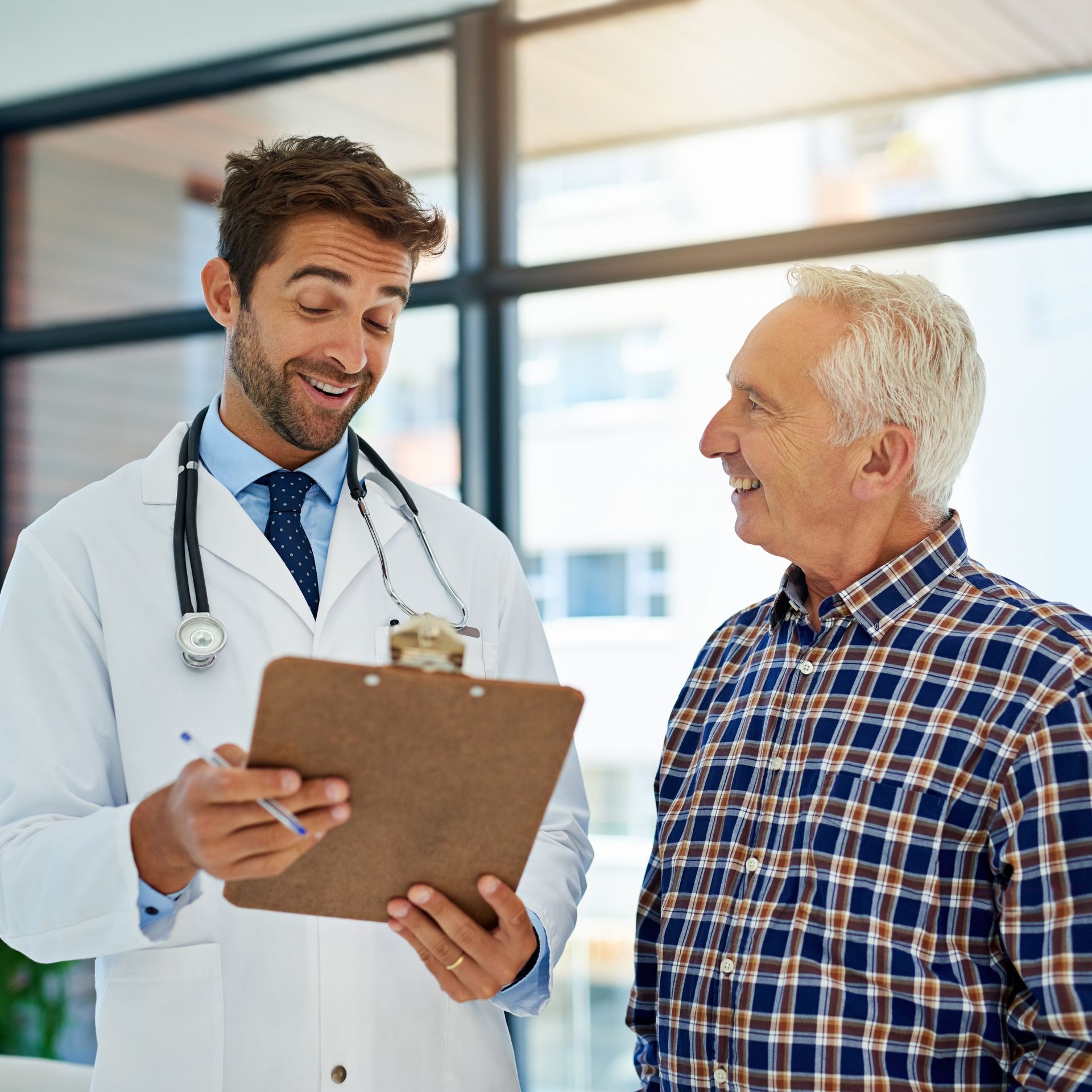 Doctor with clipboard smiles at patient, in front of a window.