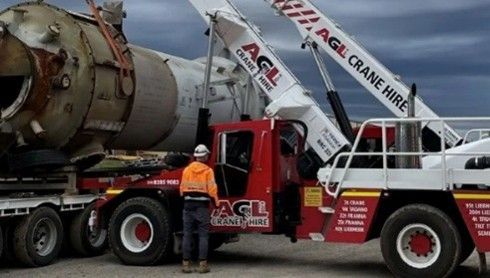 A crane labelled AGL Crane Hire lifts a large, rusted industrial tank onto a flatbed truck.