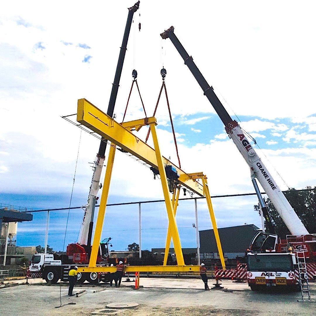Two cranes lift a large yellow gantry. Workers and vehicles are present. Outdoors.