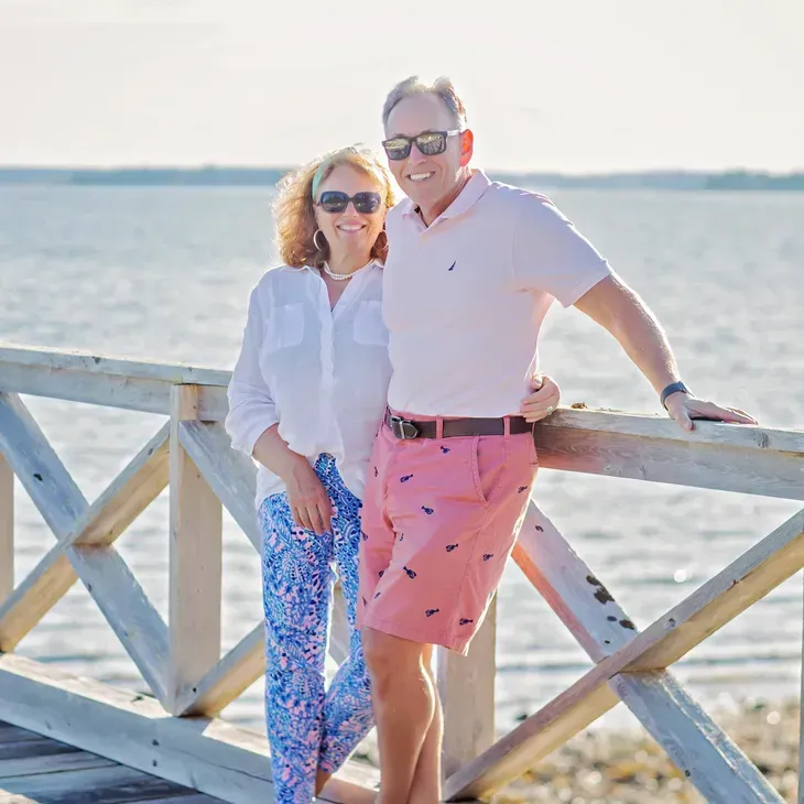 Couple smiling on a wooden pier by the water. Man in pink shorts, woman in white shirt and blue pants.