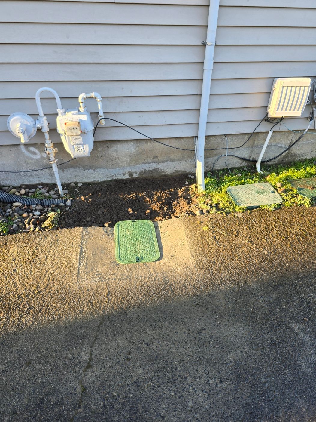 Utility meters and pipes beside a house wall, with a sidewalk and curb in front.