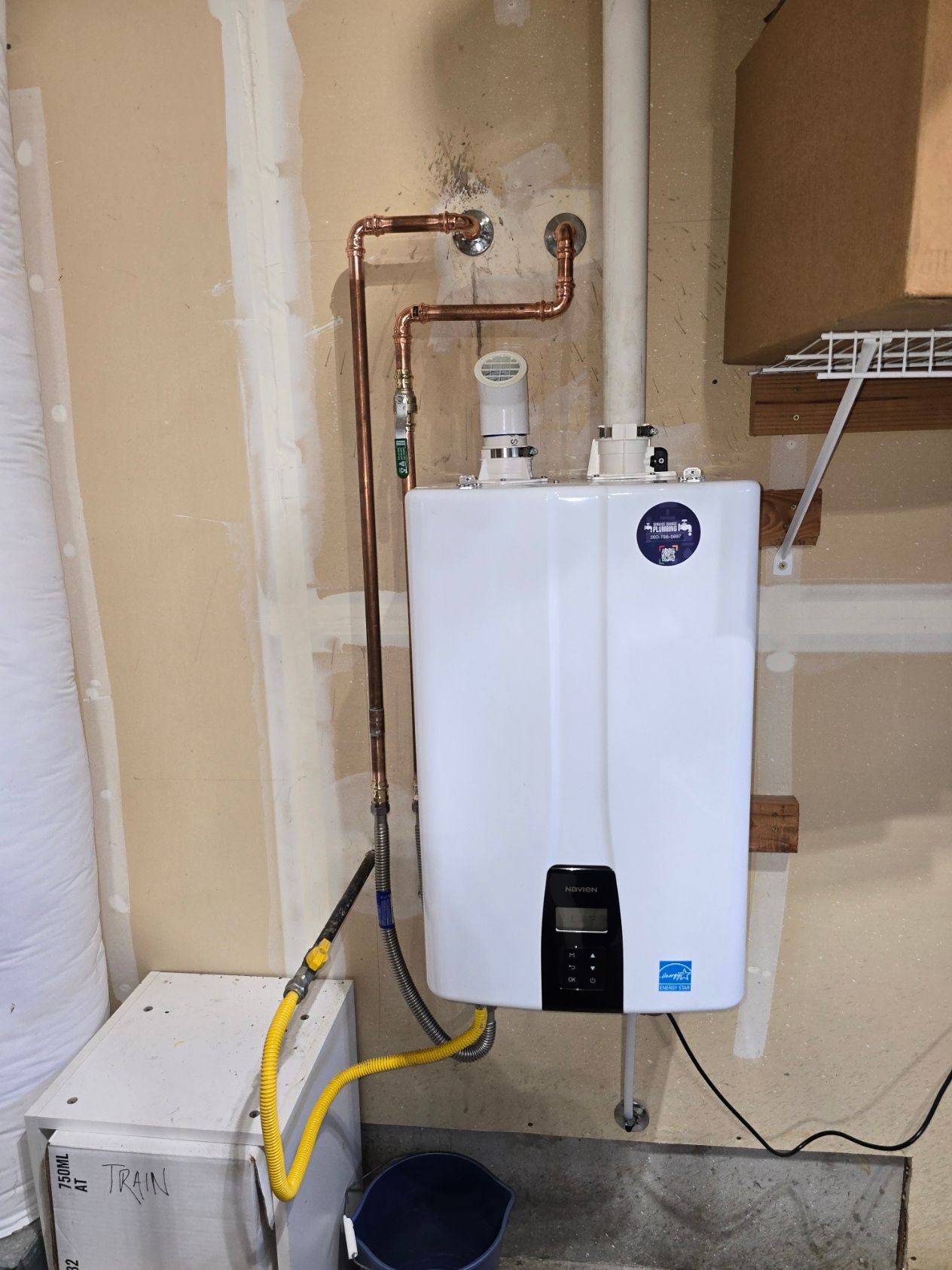 A wall-mounted white tankless water heater in a garage, connected to copper water pipes, a gas line, and white exhaust vents.