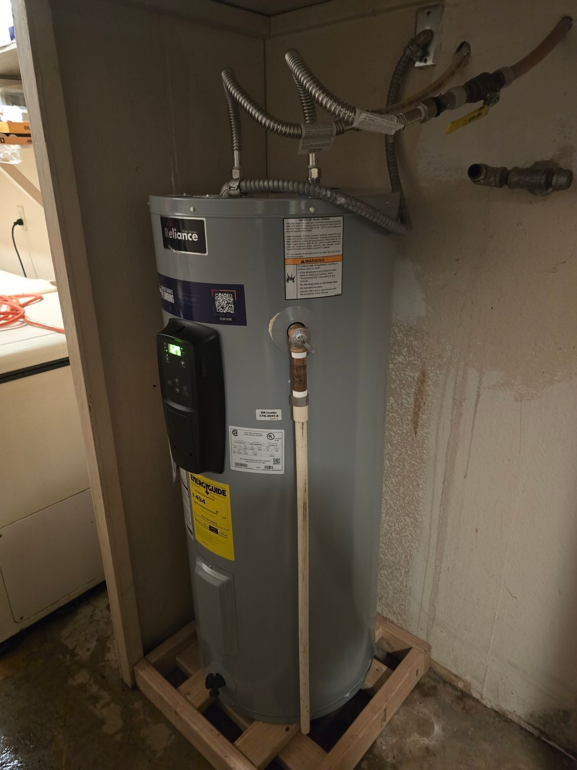Gray water heater in a utility closet with pipes and venting, set on a wooden frame against a wall.