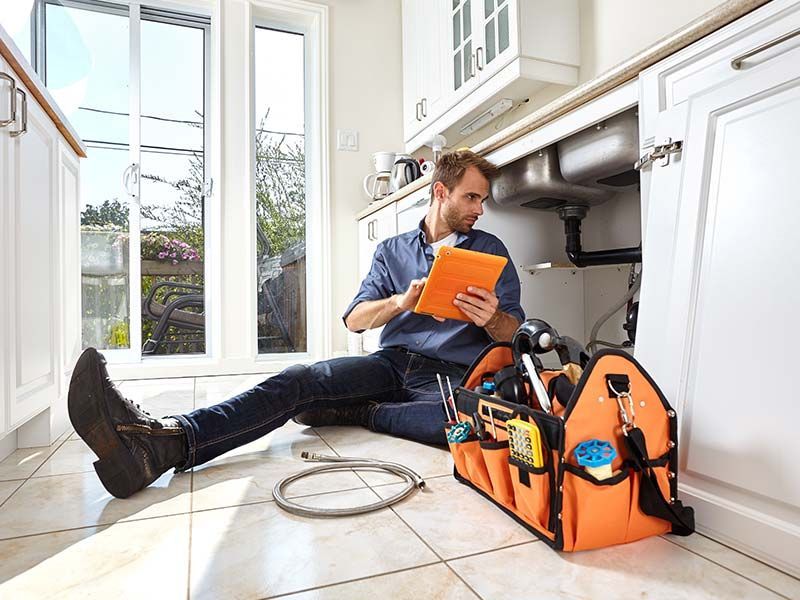 Technician seated on kitchen floor beside orange tool bag, inspecting paperwork near cabinets and a window
