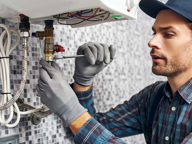Plumber tightening a pipe under a sink with a wrench in a tiled bathroom or kitchen.