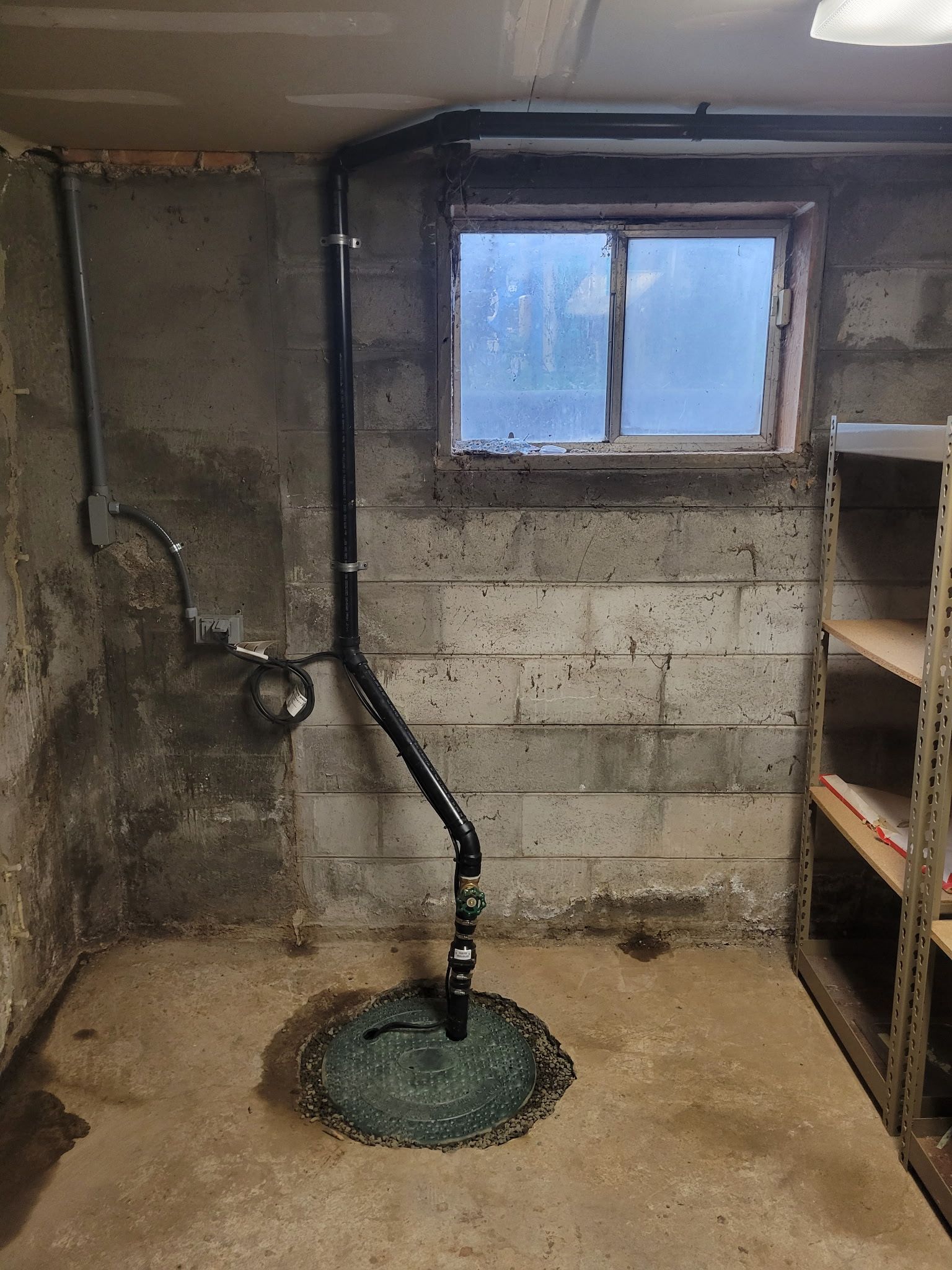Basement utility room with window, concrete walls, exposed pipes, and a floor drain with green cover