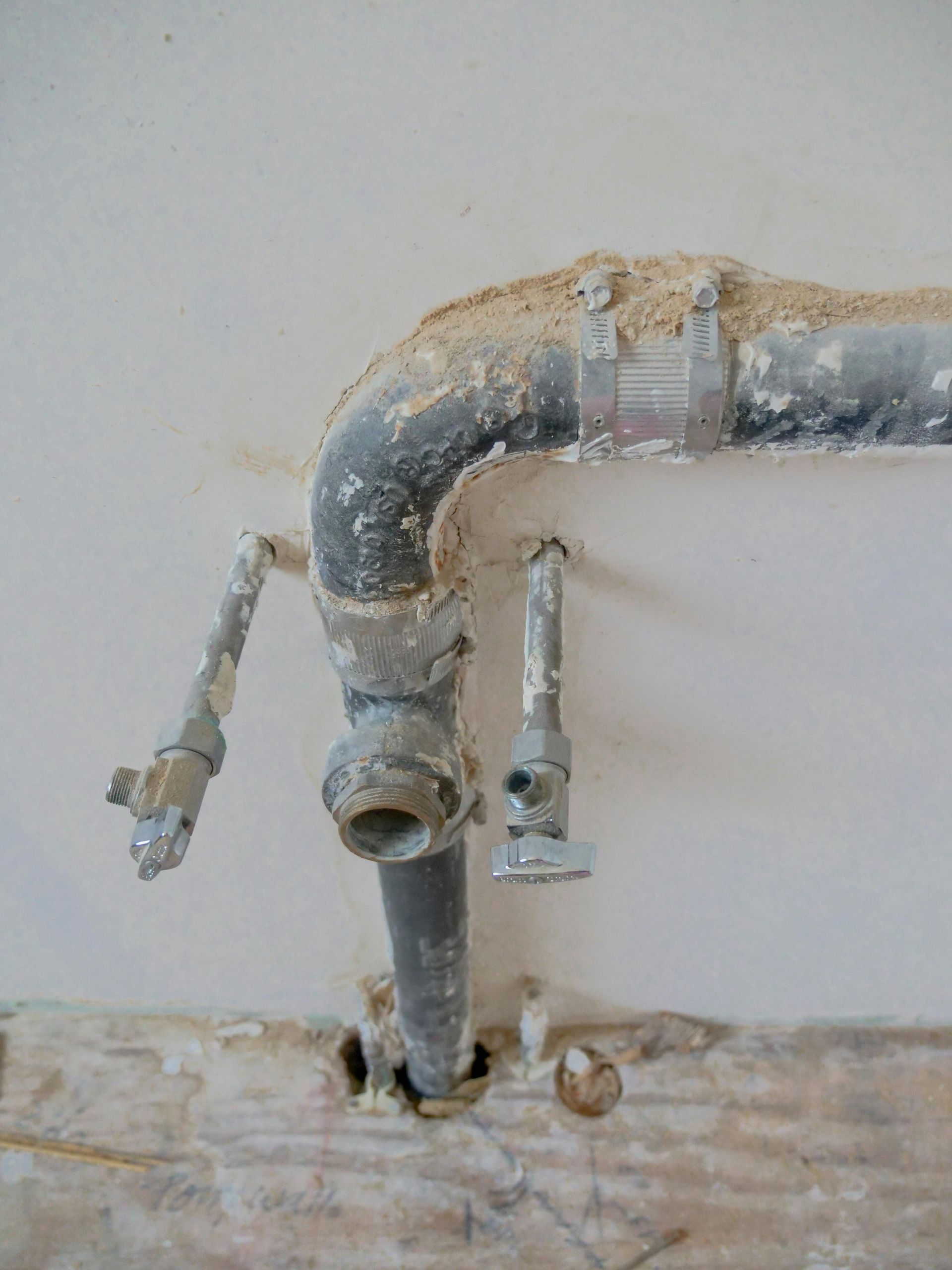 Rusty wall pipe with two small valves protruding from a damaged plaster wall.