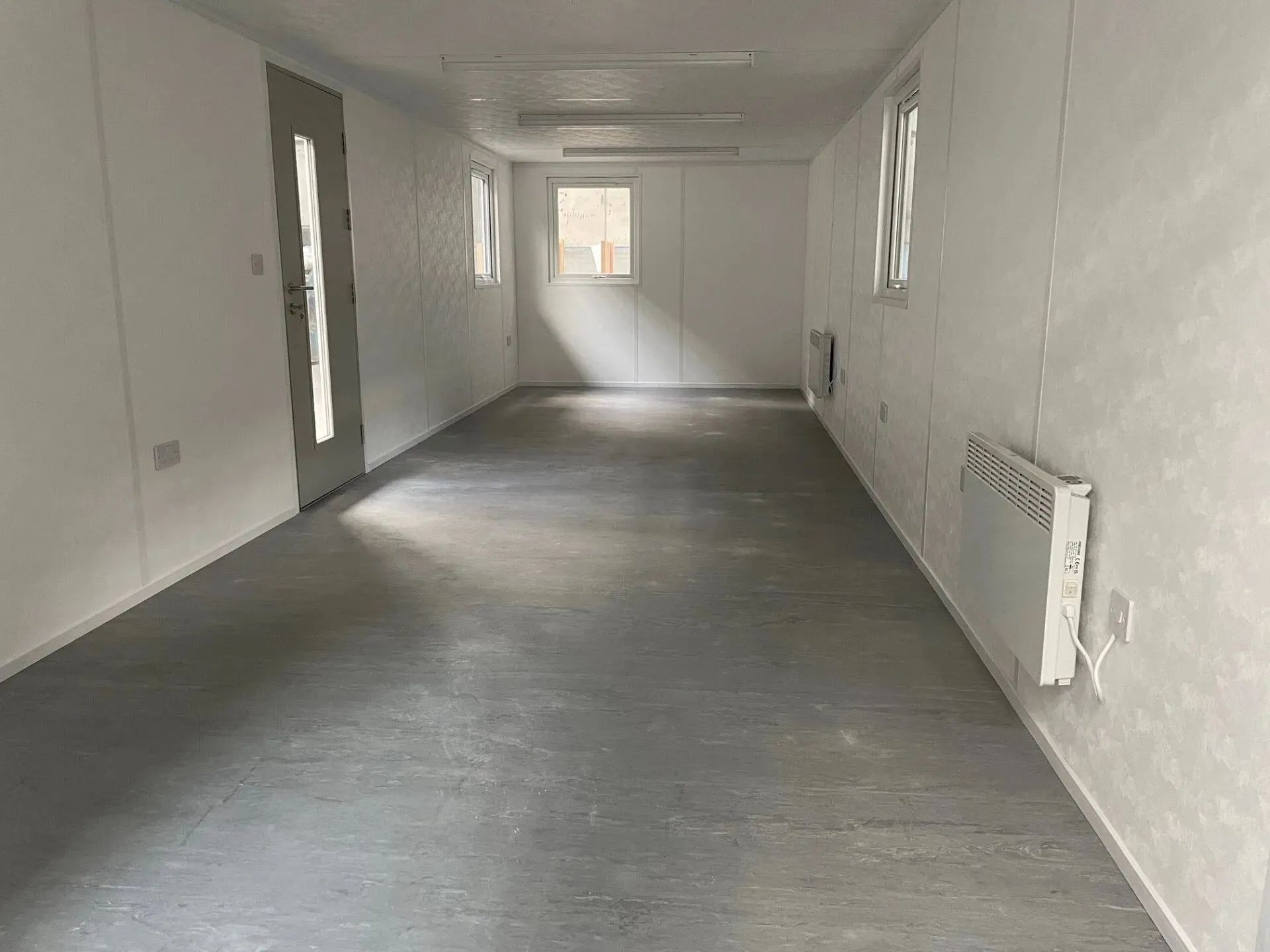 Empty portakabin interior with a door, windows, and visible radiator