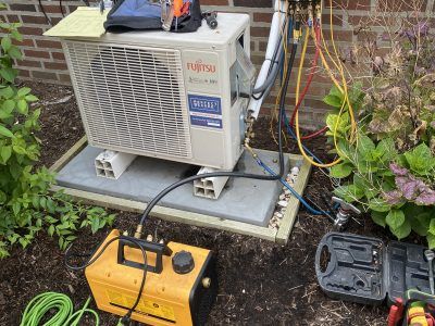 HVAC technician servicing a Fujitsu heat pump unit outdoors, with tools and gauges connected.