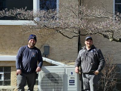 Two men standing by an air conditioning unit outside a building, posing for a photo.