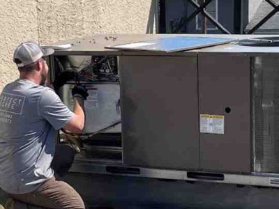 HVAC technician working on a rooftop unit. He is wearing a grey t-shirt, baseball cap, and gloves.