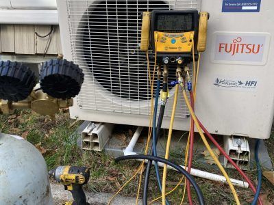 HVAC technician servicing a Fujitsu air conditioning unit outside. Yellow and black gauges connected with hoses.