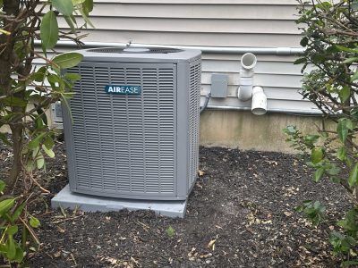 An air conditioning unit outdoors on a concrete pad, next to a house with shrubs.