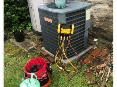 HVAC technician servicing an air conditioning unit outside. Gauges, hoses, and refrigerant tank visible.