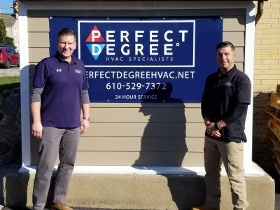 Two men stand in front of a sign for Perfect Degree HVAC. The sign is blue and white with company info.