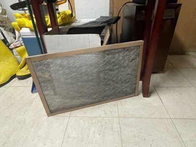Dirty air filter in a wooden frame on a tiled floor, near clutter and furniture.