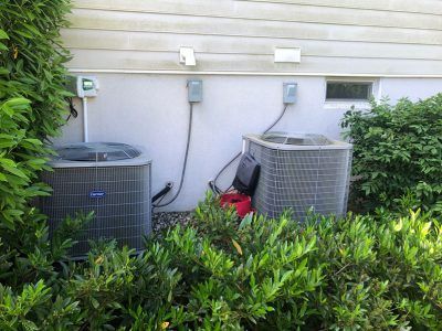 Two air conditioning units near a wall with electrical boxes and shrubbery in front.