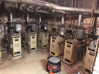 Row of beige boilers with silver pipes in a basement.