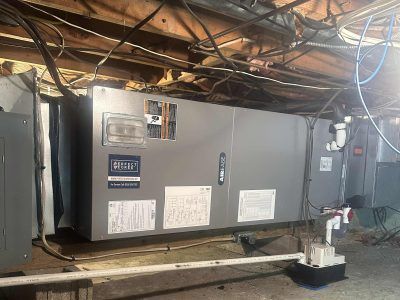 HVAC unit in a basement. The metallic unit has stickers and is surrounded by pipes, wires, and wooden beams.