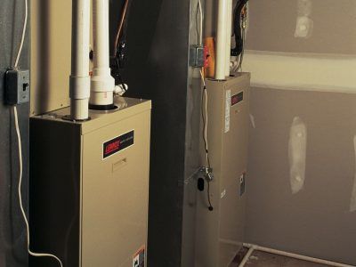 Two beige furnaces in a room with white pipes and drywall.