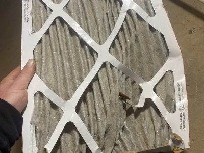 Hand holding a dirty, gray HVAC air filter with a torn cardboard frame.
