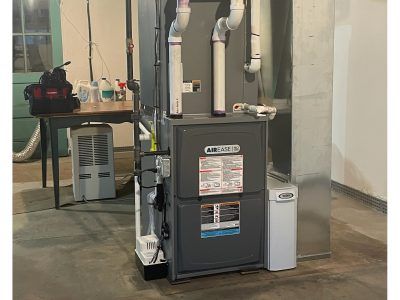 A furnace in a basement setting with white venting pipes. A table with tools is in the background.