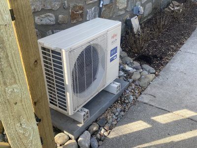 Outdoor AC unit next to a building on a concrete pad, surrounded by rocks and a wooden post.