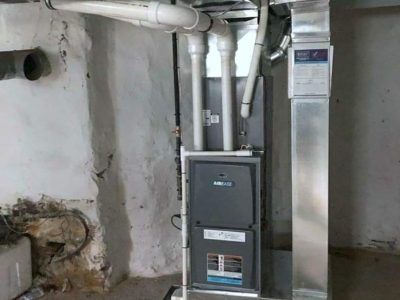 Furnace in a basement with white pipes, a metal flue, and a moldy wall.