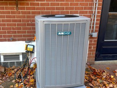 AirEase air conditioning unit outside brick building. Gray metal, fan on top, and fallen leaves.