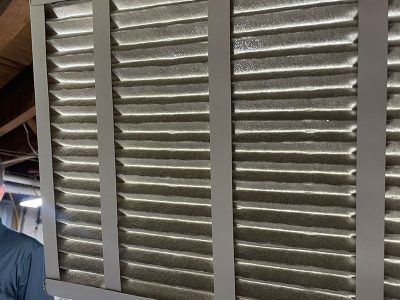 A dirty air filter in a silver frame, likely for an HVAC system, is held up against a concrete wall.