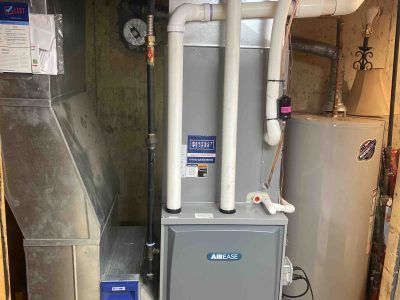 Furnace and water heater in a basement. Grey furnace, white pipes, and a silver water heater.