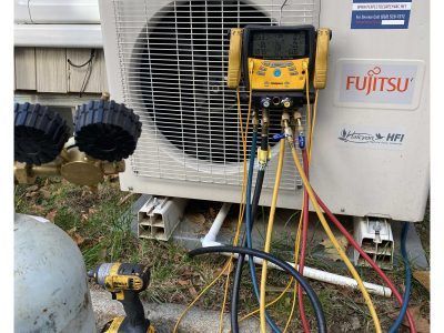 HVAC technician servicing a Fujitsu air conditioning unit outside. Gauges and hoses attached, tools nearby.