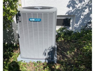 AirEase AC unit outdoors, gray metal, positioned near a white building with small window, surrounded by green foliage.