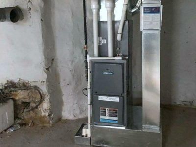 Furnace unit in a basement, with flue pipes. Gray and silver, against a white and soiled wall.