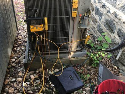 HVAC unit with gauges and hoses connected, outdoors next to stone wall and foliage.